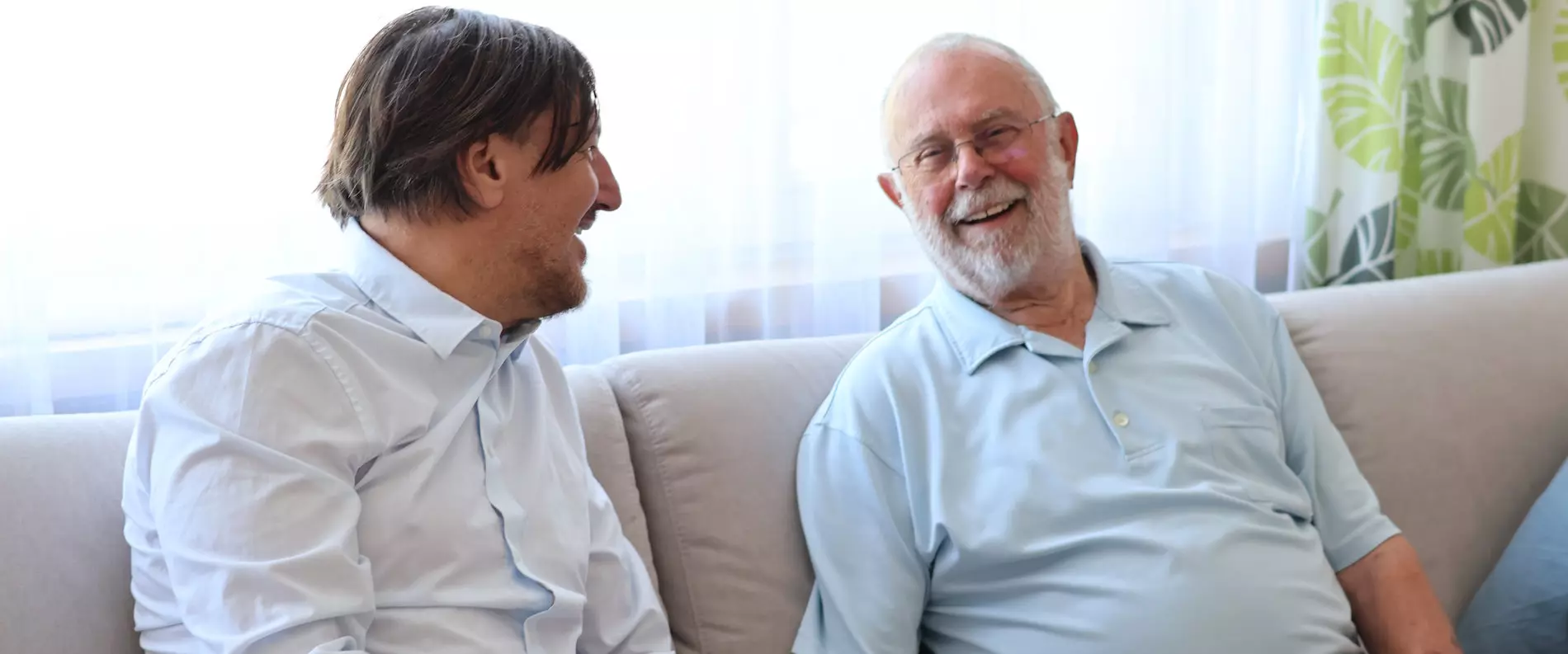 Zwei Senioren sitzen lachend und vertraut zusammen auf dem Sofa im hellen Wohnzimmer